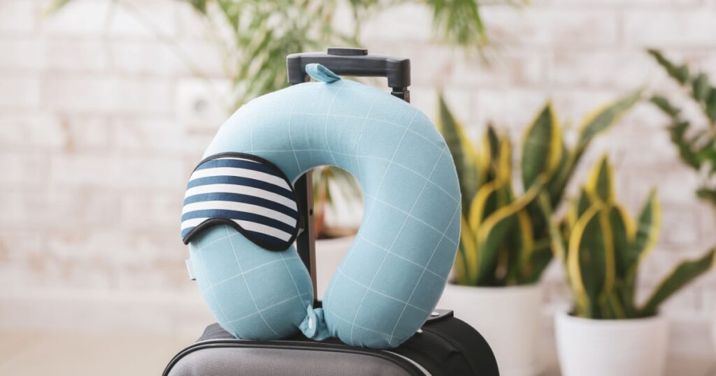 coussin de voyage bleu avec un bandeau de nuit pour les yeux déposé au-dessus d'une valise à roulette