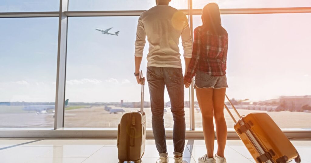 couple se tenant la main avec leur valise à l'aéroport en train de regarder les avions décoller