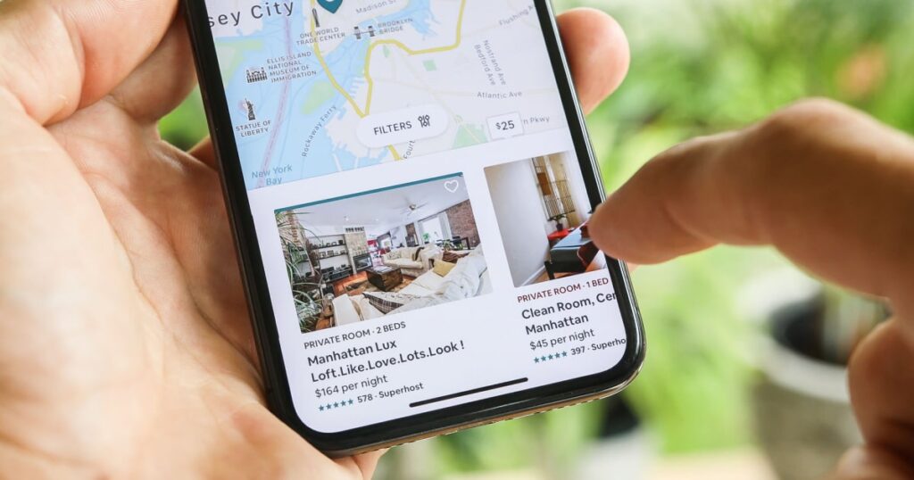 homme tenant dans ses mains son smartphone ouvert sur l'application airbnb en vue de réserver son prochain logement pour un voyage à Manhattan