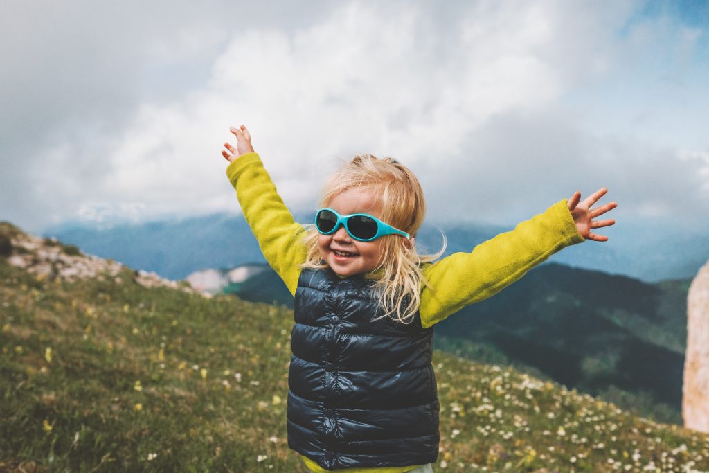 petite fille souriante avec des lunettes de solelil turquoise en vacances dans les montagnes en train de lever les bras