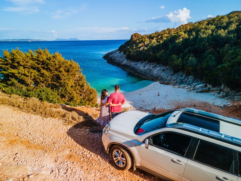 couple en voyange se serrant les bras devant leur voiture de location en regardant une plage de sable fin